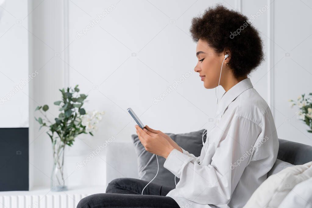 side view of pleased african american woman listening music and using smartphone in living room