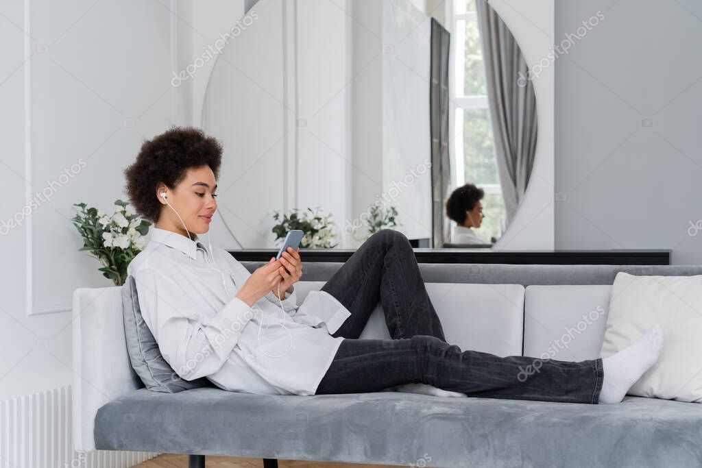 Joyful african american woman listening music in wired earphones and using smartphone in modern living room