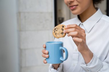 cropped view of happy african american woman holding cup of milk and cookie with chocolate chips  clipart