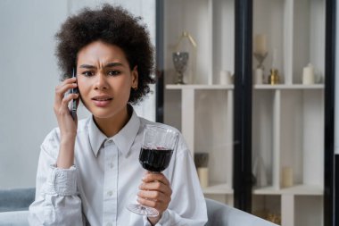 Üzgün Afrikalı Amerikalı kadın elinde bir bardak kırmızı şarap tutarak akıllı telefondan konuşuyor. 