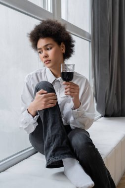 sad african american woman holding glass of red wine and looking at window while sitting at windowsill  clipart