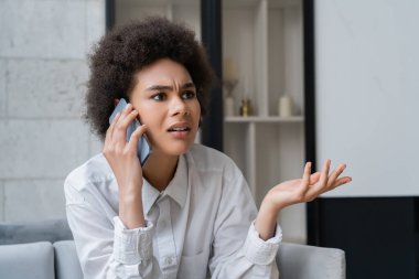Beyaz gömlekli, hoşnutsuz Afro-Amerikan kadın akıllı telefondan konuşuyor ve evde el kol hareketi yapıyor.
