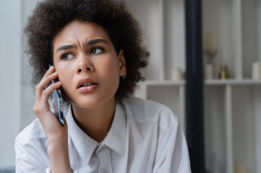 Beyaz gömlekli Afrikalı Amerikalı kadın evde akıllı telefondan konuşuyor.