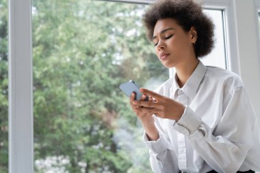 Beyaz tişörtlü, üzgün Afro-Amerikan bir kadının pencerenin yanındaki akıllı telefondan mesaj okuması.
