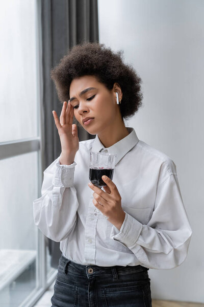 sad african american woman in white shirt and wireless earphone holding glass of red wine 
