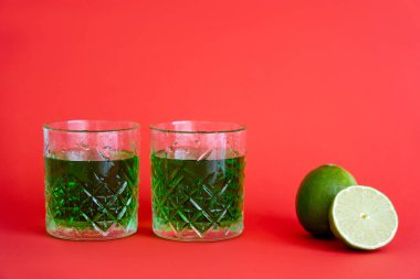 green alcohol drink in faceted glasses with water drops near limes on red clipart