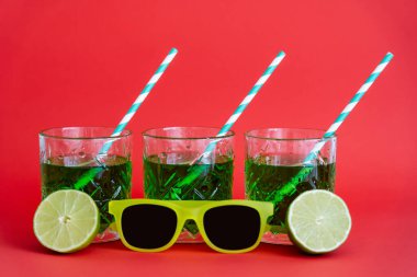 Kırmızı gözlüklü güneş gözlüğü yanında kamış ve yarım dilim limon içeren yeşil bardakta içki.