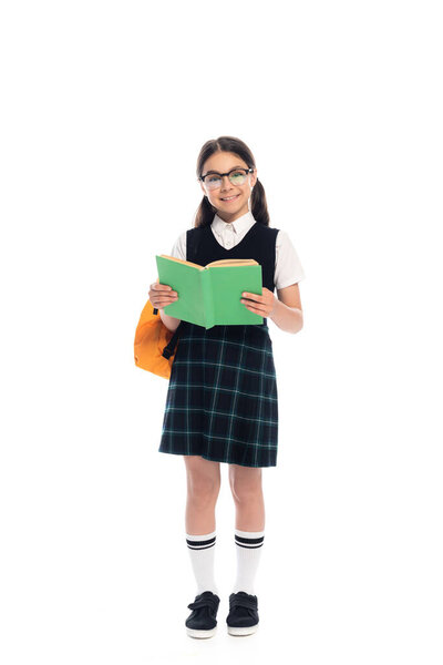 Full length of schoolkid in eyeglasses holding book and smiling at camera on white background