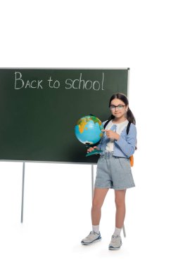 Gülümseyen okul çocuğu yazı tahtasının yanındaki küreyi işaret ediyor ve beyaz arka planda okula geri dönüyor.