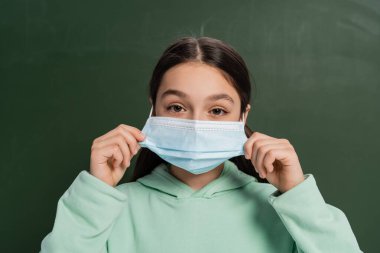 Esmer okul çocuğu arka planda karatahtanın yanında tıbbi maske takıyor.