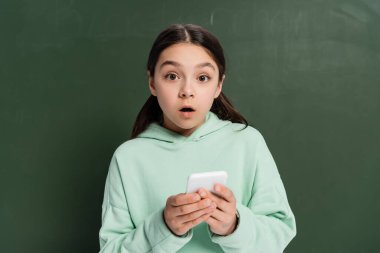 Şok olmuş okul çocuğu arka planda yazı tahtasının yanında akıllı telefon tutuyor.