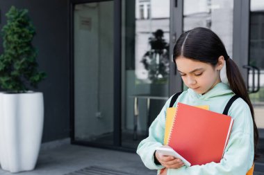 Not defterleri olan liseli kız şehir caddesinde akıllı telefon kullanıyor. 