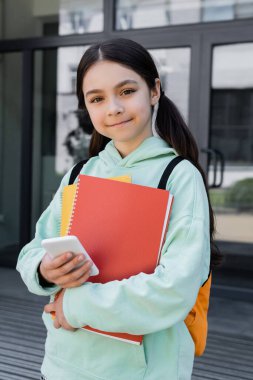 Elinde cep telefonu ve defter tutan gülümseyen okul çocuğu. 
