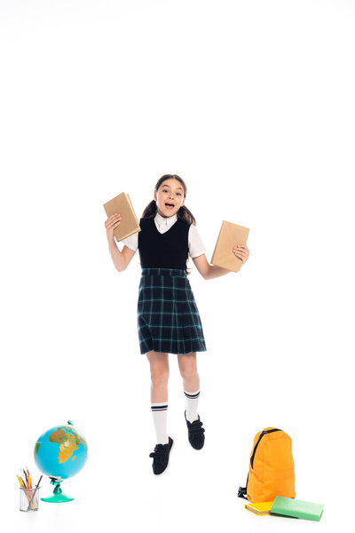 Excited schoolkid holding books while jumping near globe and backpack on white background
