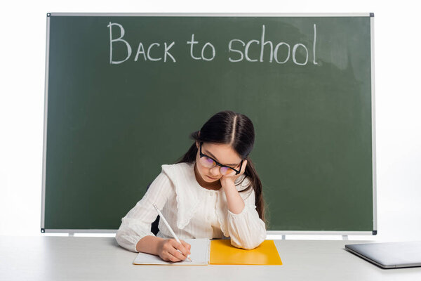 Pupil in eyeglasses writing on notebook near laptop and chalkboard with back to school lettering isolated on white 