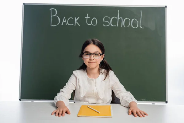 Smiling schoolgirl looking at camera near notebook and chalkboard with back to school lettering isolated on white