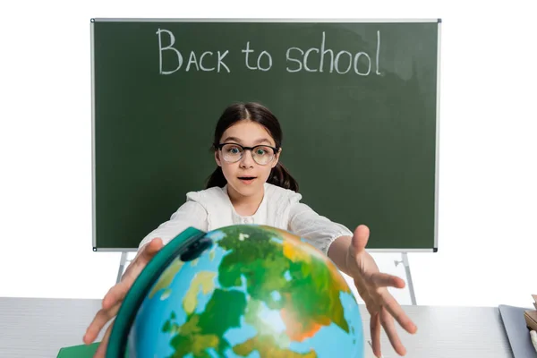 Excited schoolgirl outstretching hands to globe near chalkboard with back to school lettering isolated on white