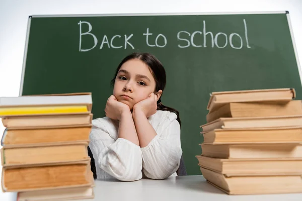 Schoolkid looking at camera near blurred books and chalkboard with back to school lettering isolated on white