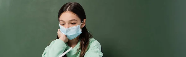 Preteen schoolgirl in medical mask holding pencil near chalkboard at background, banner