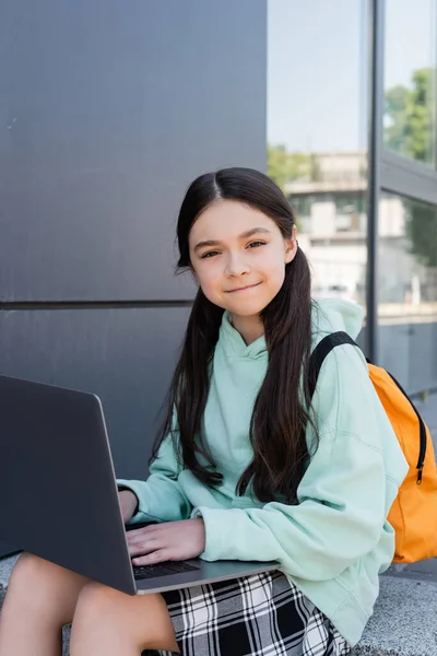 Positive schoolgirl with backpack using laptop near building outdoors