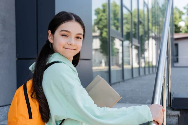 Positive pupil with backpack and books looking at camera near blurred books outdoors