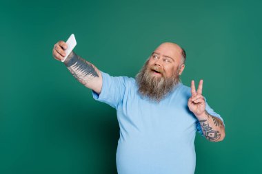 happy and bearded overweight man showing victory sign while taking selfie isolated on green clipart