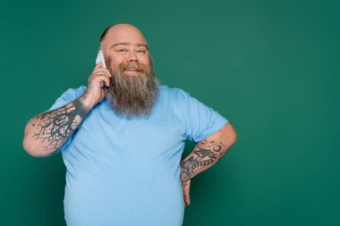 happy and bearded plus size man standing with hand on waist and talking on cellphone isolated on green