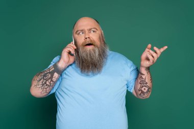 thoughtful overweight man with tattoos talking on smartphone and pointing with finger isolated on green
