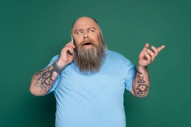 thoughtful overweight man with tattoos talking on smartphone and pointing with finger isolated on green clipart