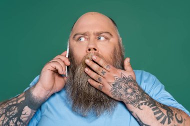 shocked tattooed man covering mouth with hand while talking on smartphone isolated on green