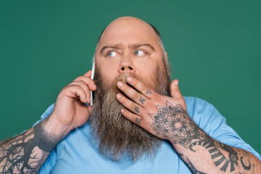 shocked tattooed man covering mouth with hand while talking on smartphone isolated on green clipart