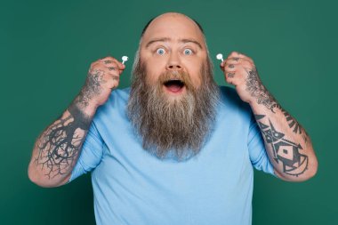 amazed chubby man with beard holding wireless earphones isolated on green