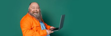 cheerful and bearded plus size man with laptop looking at camera isolated on green, banner