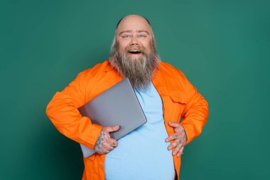 pleased overweight man with laptop touching tummy isolated on green