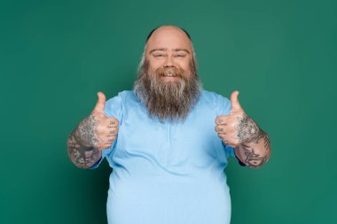 bearded overweight man showing thumbs up and smiling at camera isolated on green