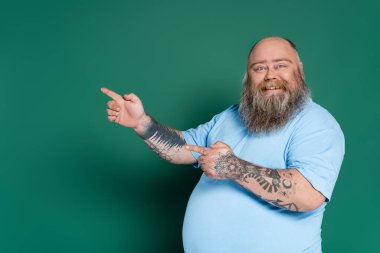 cheerful plus size man with beard pointing with fingers isolated on green