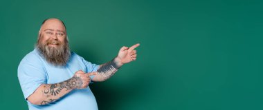 cheerful and chubby man pointing with fingers isolated on green, banner