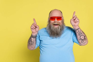 bearded and cheerful overweight man in sunglasses pointing up with fingers isolated on yellow clipart