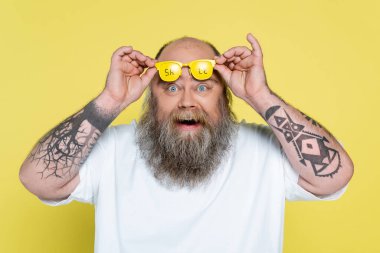 amazed tattooed plus size man holding glasses with sale lettering isolated on yellow