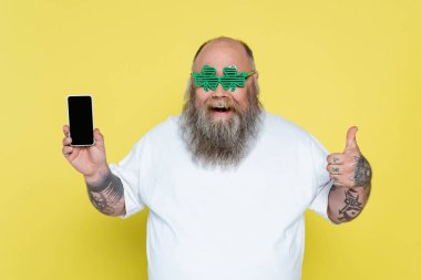 joyful overweight man in clover-shaped glasses showing thumb up while holding smartphone isolated on yellow