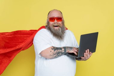 thrilled overweight man in red sunglasses and superhero cloak using laptop isolated on yellow clipart