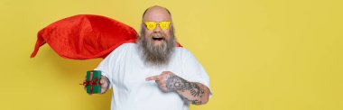 excited plus size man in red cloak and sunglasses with sale lettering pointing at gift box isolated on yellow, banner