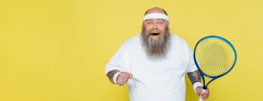 excited overweight man with tennis racquet looking at camera isolated on yellow, banner