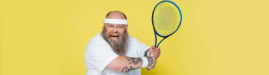 thrilled plus size man with beard and tennis racquet looking at camera isolated on yellow, banner