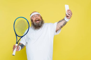happy and chubby man taking selfie with tennis racquet isolated on yellow