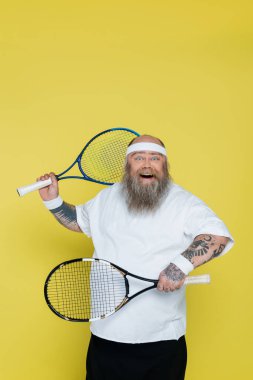 cheerful overweight tennis player with racquets looking at camera isolated on yellow