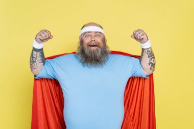 bald and bearded overweight sportsman in superhero cloak showing power isolated on yellow