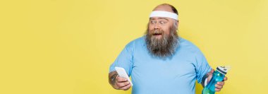 amazed plus size man with sports bottle looking at smartphone isolated on yellow, banner