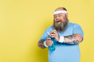 thirsty overweight man with beard opening sports bottle isolated on yellow