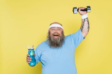 happy overweight man holding sports bottle while training with dumbbell isolated on yellow clipart